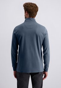 Uomo con capelli ricci castani che indossa una camicia blu a maniche lunghe e pantaloni grigio scuro, in piedi di fronte a uno sfondo grigio chiaro uniforme.