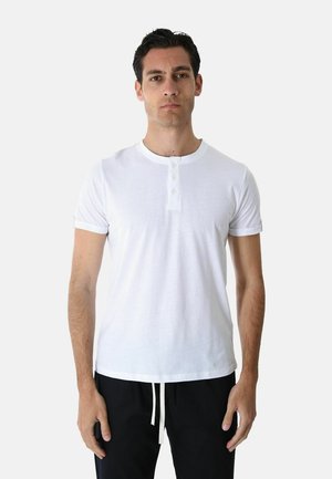 Uomo che indossa una maglietta henley bianca semplice a maniche corte e pantaloni neri, in piedi di fronte a uno sfondo grigio chiaro.