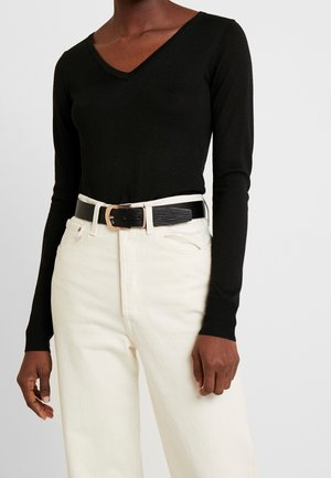 Personne portant un haut noir à manches longues et col en V rentré dans un pantalon blanc taille haute avec une ceinture noire à boucle dorée.