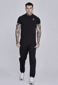 Černé tričko s krátkým rukávem s kontrastním logem na rukávech a černé tepláky. Model má na sobě bílé sportovní boty.