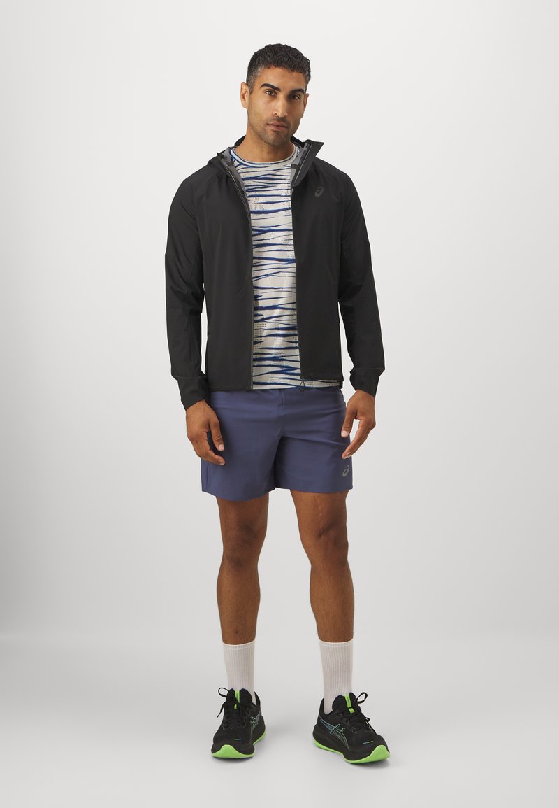 Chaqueta ligera negra, camisa a rayas azul y blanca, pantalones cortos azul marino y zapatillas de correr negras con acentos verdes. Medias deportivas blancas visibles.