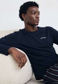 Marine langærmet T-shirt med broderet "HILFIGER" logo på brystet. Model siddende, tekstureret baggrund med en synlig hånd hvilende på en blød overflade.