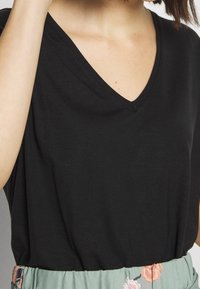 Svart v-ringad t-shirt i mjukt tyg, med korta ärmar och avslappnad passform, ihopparad med ett blomstermönstrat grönt midjeband.