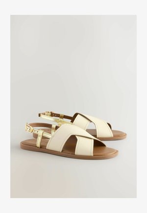 Jasne sandały skórzane w kolorze kremowym, z otwartym noskiem i wyciętymi detalami. Regulowane zapięcia w odcieniu złota; płaska brązowa podeszwa.