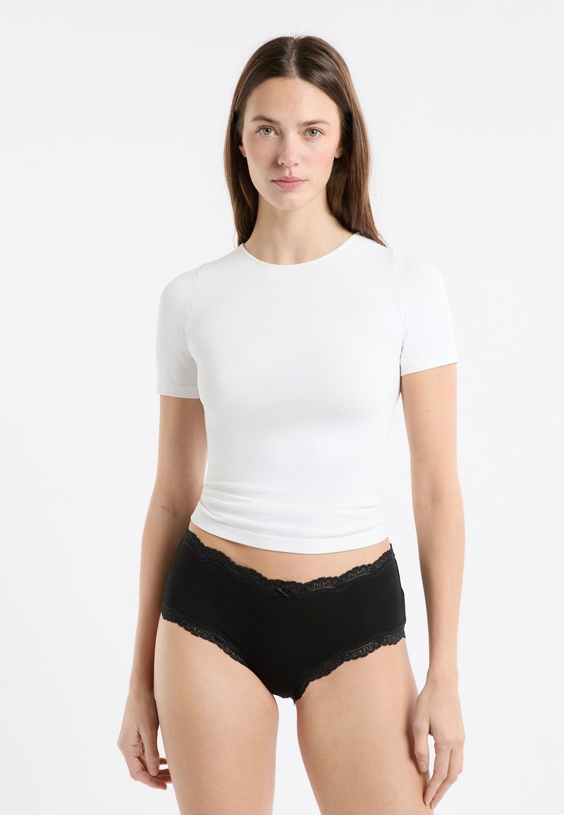 Weißes Kurzarmshirt mit rundem Ausschnitt kombiniert mit schwarzer hoch taillierter Unterwäsche, die am Bund mit Spitzenbesatz verziert ist.
