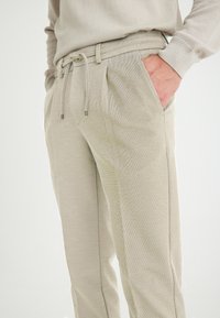 Lichtbeige trainingsbroek van een gestructureerde stof, met een elastische tailleband, trekkoorddetails en zijzakken, voorzien van subtiele stikdetails.