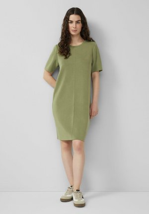 Frau mit langen, lockigen Haaren, die ein knielanges, olivgrünes Kurzarmkleid und beige Turnschuhe trägt, steht vor einer hellgrauen Wand.