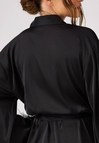 Czarna satynowa szlafrok z szerokimi rękawami, przewiązany w talii, o gładkiej fakturze i delikatnym połysku. Tylny widok podkreśla design kołnierza.