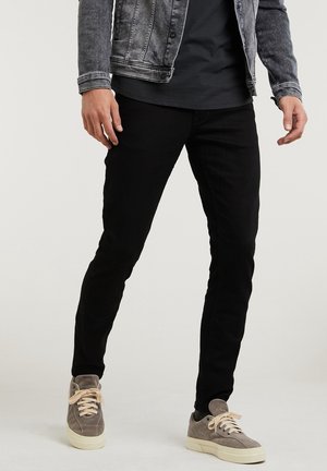 Uomo che indossa pantaloni neri aderenti, sneakers grigie in suede, t-shirt nera e giacca di jeans grigia in piedi contro uno sfondo uniforme.