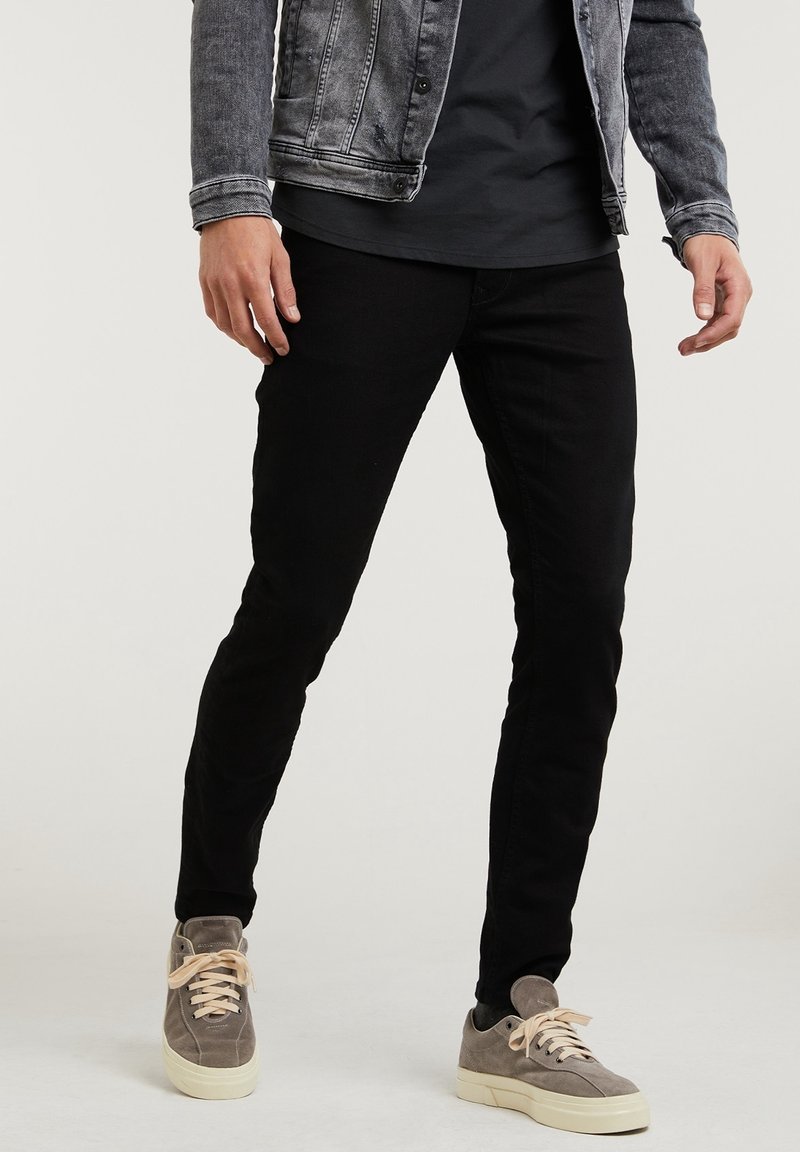 Homme portant un pantalon noir ajusté, des baskets en daim gris, un t-shirt noir et une veste en denim grise, debout devant un fond uni.