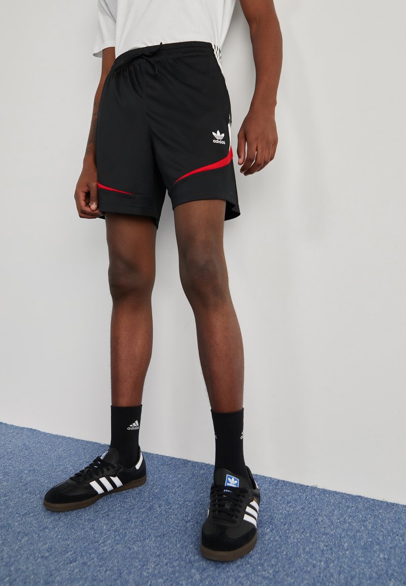 Černé sportovní kraťasy s červenými akcenty a logem Adidas, se elastickým pasem. Nosič má na sobě černé boty a ponožky délky do poloviny lýtek.