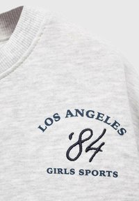 Sivá farebná mikina s rebrovaným výstrihom. Obsahuje námornícke výšivky: "LOS ANGELES '84 GIRLS SPORTS" v okrúhlom dizajne.