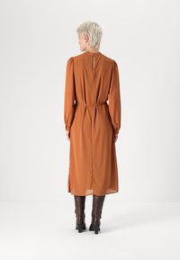 Rochie lungă de culoare portocalie închisă, cu mâneci lungi, talie strânsă, umeri plisați, închidere cu butoane la spate și fante laterale, asortată cu cizme maro până la genunchi.