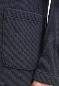 Tessuto blu navy con un motivo a nido d'ape, caratterizzato da una tasca applicata e cuciture grezze sulla manica. Design casual e dal taglio rilassato.