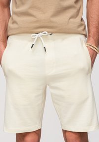 Homme portant un short beige à cordon de serrage et un t-shirt beige, les mains dans les poches et des bracelets de perles au poignet droit.
