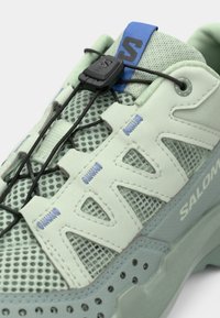 Lichtgroene mesh en synthetische sneaker met zwart snelvetersysteem, blauw tonglabel en "Salomon" branding aan de zijkant van de zool.