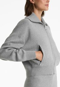 Vrouw in een lichtgrijze zip-up trui met hoge kraag, met handen deels in de gezuipte zijzakken, zijwaarts en vanaf de borst omhoog afgebeeld.