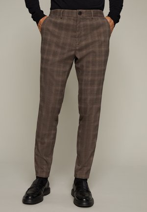 Herren braune karierte maßgeschneiderte Hose, getragen mit schwarzen Schuhen und Händen in den Taschen vor einfachem Hintergrund.