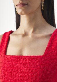 Punainen teksturoitu mekko, jossa on neliön muotoinen pääntie ja leveät hihat. Pystyraita-muotoilu, yhdistettynä kultaisiin roikkuviin korvakoruihin, joissa on helmiä.