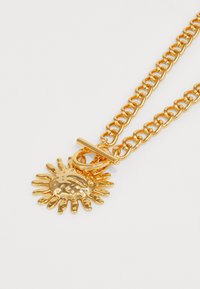 Colar de metal dourado com um grande pendente em forma de sol com raios. Corrente de elos grossos com fecho de encaixe para um fechamento seguro.
