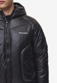 Svart pufferjacka med dragkedja fram, huva och logotyp. Har en quiltad design, slät textur och dragkedjefickor på sidorna.