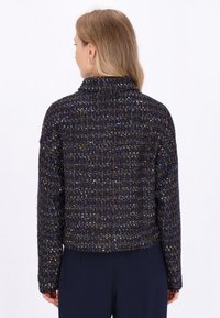 Una donna con lunghi capelli biondi indossa una giacca blu navy con una texture e macchie colorate, su uno sfondo bianco uniforme.