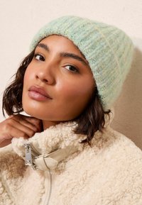 Gaiši zils adīts cepure ar teksturētu virsmu, apvienota ar krējuma krāsas, pūkainu, ar rāvējslēdzēju, jaku. Tuvplāns uz sejas iezīmēm.