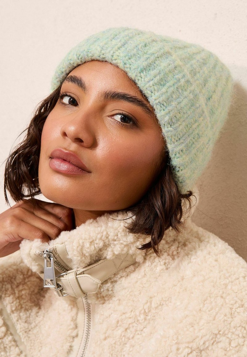 Gaiši zils adīts cepure ar teksturētu virsmu, apvienota ar krējuma krāsas, pūkainu, ar rāvējslēdzēju, jaku. Tuvplāns uz sejas iezīmēm.