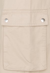 Tissu beige avec une poche équipée de boutons-pression argentés. La poche est de forme carrée avec des surpiqûres détaillées autour des bords.