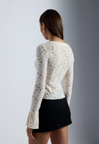 Lace-Oberteil in Elfenbein mit floralem Muster, langen Ärmeln und ausgestellten Manschetten, kombiniert mit einem schwarzen Rock. Strukturiertes, halbtransparentes Design.