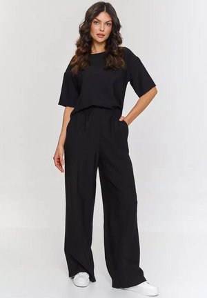 Kobieta z długimi falującymi włosami, ubrana w luźną czarną bluzkę z krótkim rękawem i szerokie spodnie oraz białe trampki, stojąca z jedną ręką w kieszeni.