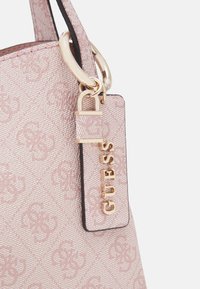 Rosa mönstrad handväska med texturerat tyg, guldfärgade detaljer och en etikett med "GUESS" i guldbokstäver fäst med en guldring.