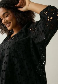 Schwarze Bluse mit floralem Lochmuster, lockere Passform und elastischen Bündchen. Mit V-Ausschnitt und strukturiertem Stoff.