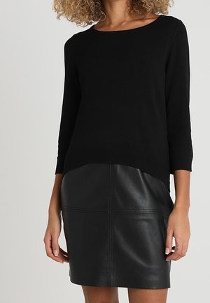 Woman wearing a black three-quarter sleeve top paired with a black leather skirt, standing against a plain light background.