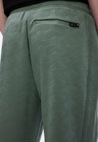 Grüne texturierte Hose mit geometrischem Muster, elastischem Bund und einer mit Reißverschluss versehenen Gesäßtasche.