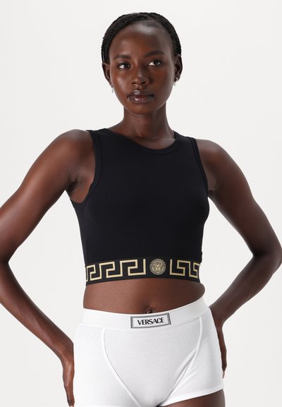 Black crop top with gold detailing featuring a Greek key pattern and logo. Paired with white ribbed boxer shorts displaying the Versace label.