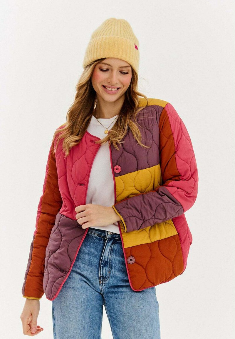 Prešívaná bunda v ružových, fialových a žltých odtieňoch. Má okrúhle gombíky, dlhé rukávy a hebký povrch. Nosená cez bielu košeľu.