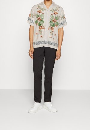 Uomo che indossa una camicia beige a maniche corte con stampa floreale, pantaloni neri e sneaker bianche, in piedi davanti a uno sfondo chiaro uniforme.