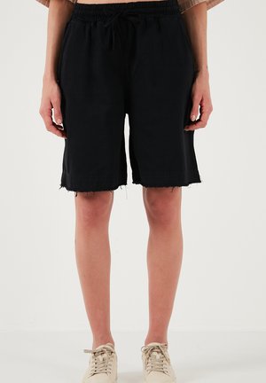 Inimene, kes kannab lahtisi musti põlvekõrguseid lühikesi pükse palja äärega ja beeže tossusid, seisab toas tavalise valge taustaga.