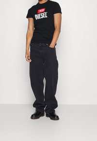 Svart Diesel t-shirt med vitt logotyp på bröstet, kombinerad med lösa svarta jeans och svarta skor med bruna sulor.