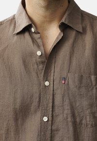 Hnědá lněná košile s límcem na knoflíky, má bílé knoflíky a kapsu na levé části hrudi. Malé logo na kapse.