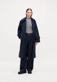 Marineblå ullfrakk med bred krage, stripete skjorte under, og løse mørke denimbukser. Svarte platåsko fullfører antrekket.