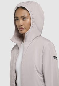 Light pink hooded jacket made of smooth, water-resistant fabric. Features a front zipper and a black logo patch on the sleeve.