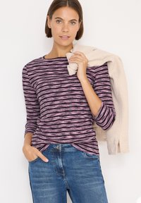 Maglietta a maniche lunghe a righe viola con righe nere e rosa, abbinata a jeans blu. Un maglione di colore chiaro è appoggiato su una spalla.