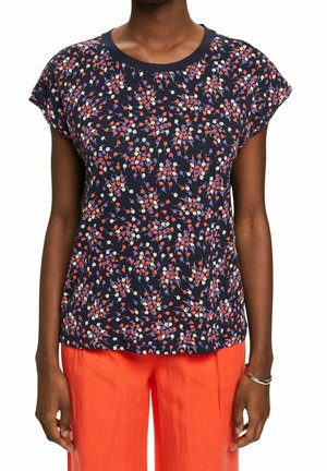 Osoba ubrana w granatową koszulkę z krótkim rękawem w czerwono-biało-fioletowy kwiatowy wzór oraz jaskrawopomarańczowe spodnie.