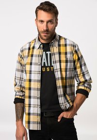 Skjorte med knaplukning i sort, hvid og gulternet mønster; lange ærmer oprullet. Båret over en sort t-shirt og sorte jeans.