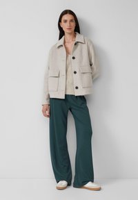 Donna in piedi contro un muro bianco, indossa una giacca beige con bottoni, un top in maglia crema, pantaloni larghi verde scuro e sneakers bianche.