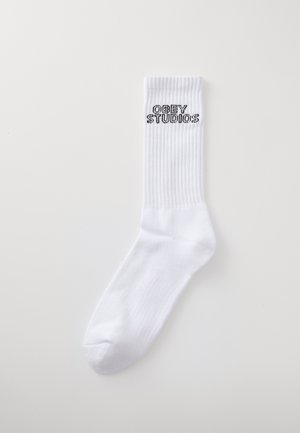 Bela bombažna nogavica z rebrastim robom, ki na zgornji strani nosi črni logotip "OBEY STUDIOS". Gladka tekstura, standardna športna oblika.