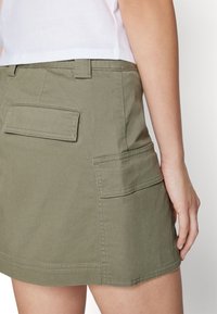 Jupe vert olive avec une coupe droite, dotée de deux poches arrière et de coutures décoratives. Fabriquée en tissu robuste avec une texture lisse.