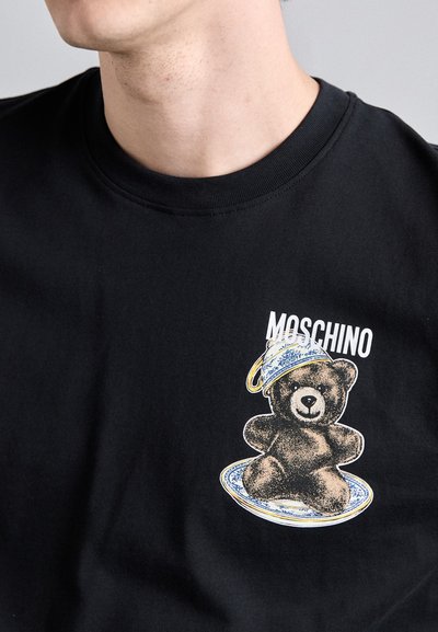 Svart bomull T-skjorte med et grafisk trykk av en bjørn som har på seg en blå-hvit mønstret hatt, med "Moschino"-tekst over illustrasjonen.